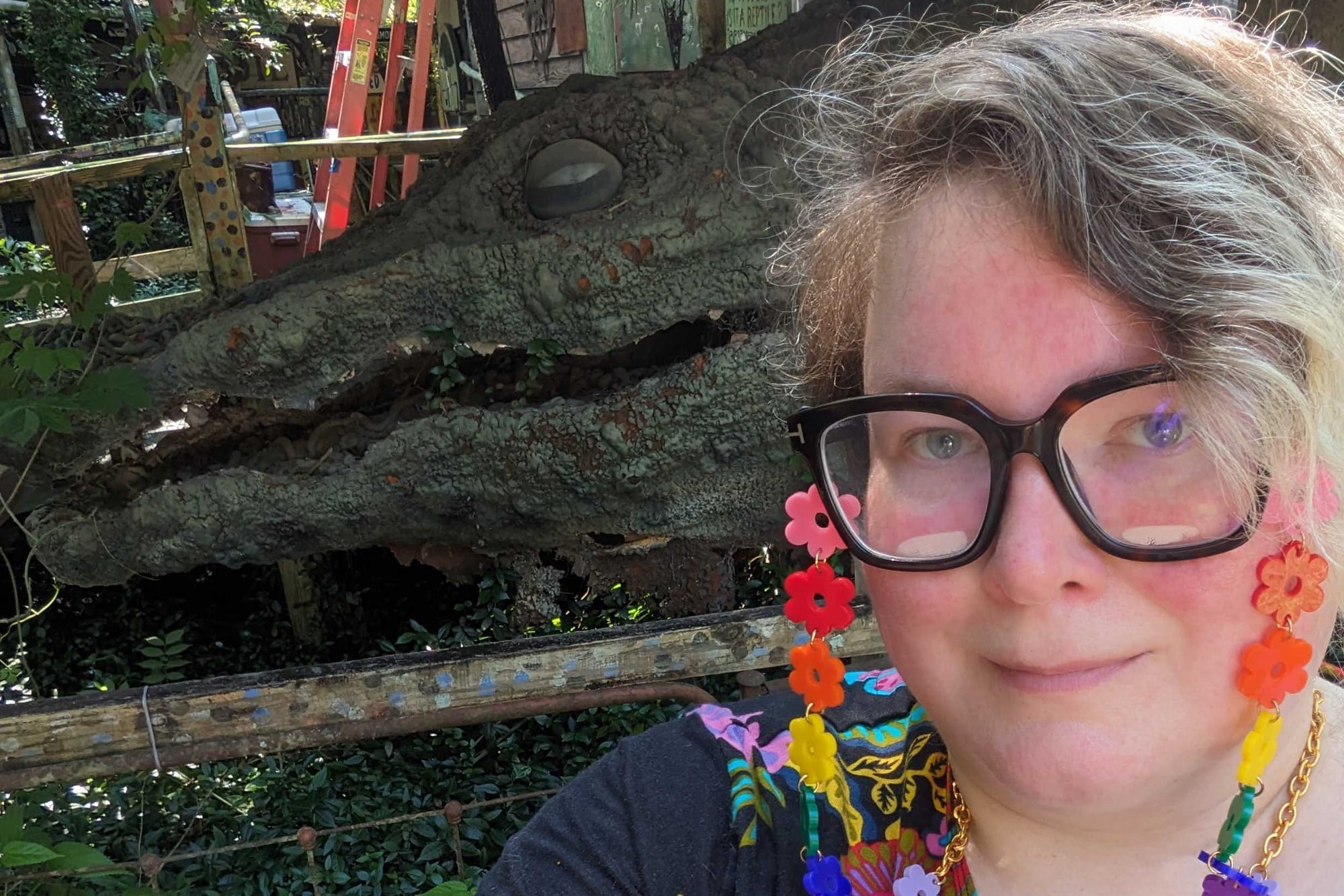 A nonbinary person with rainbow eyeglass jewelry standing in front of a giant alligator head.