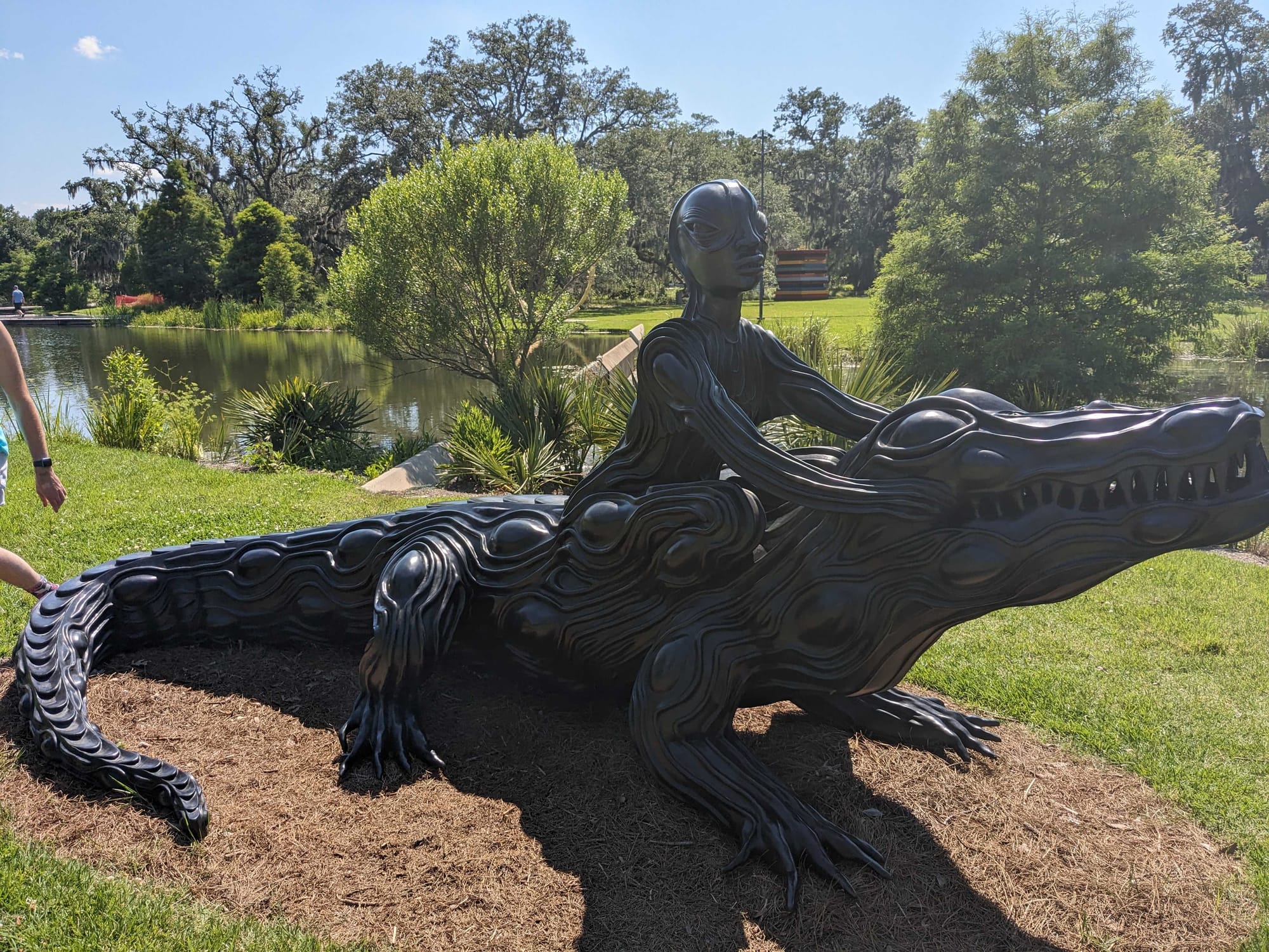 A stylized sculpture of an alien figure riding an alligator in front of the bayou.