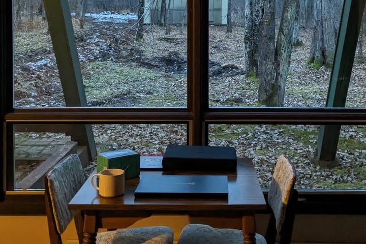 A small table set with a mug, two laptops, and some writing gear in front of a window with a winter forest view. 
