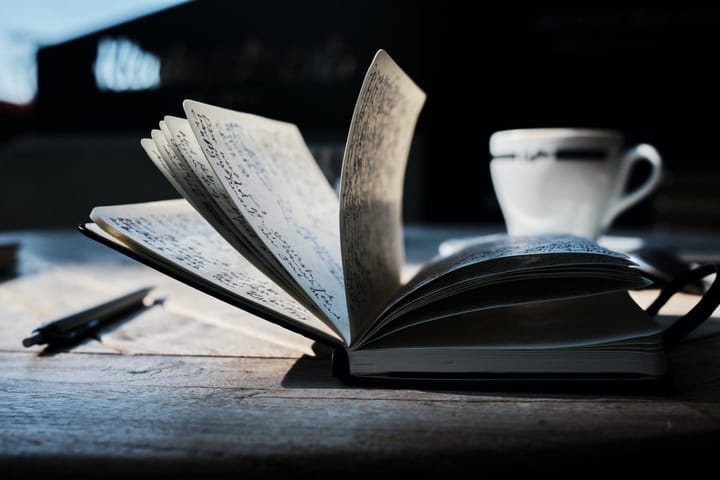 A book with fanning pages next to a pen and a coffee cup on a rough-hewn wooden table. 